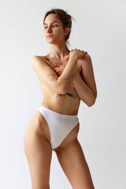Portrait of young slim girl with tattoos, covering breast with hands, posing in underwear against grey studio background. Concept of body and skin care, figure, fitness, health, wellness. - 写真・画像