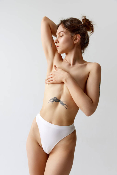 Portrait of slim young girl covering breast with hand, posing in underwear against grey studio background. Female health care. Concept of body and skin care, figure, plastic surgery, wellness. - Foto, Bild