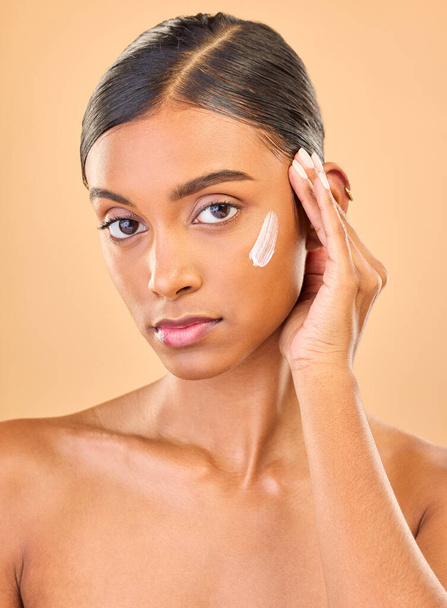 Face portrait, skincare and woman with cream in studio isolated on brown background. Dermatology, serious cosmetics and confident Indian female model with lotion, creme or moisturizer for skin health. - Φωτογραφία, εικόνα