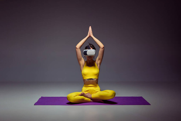 Young slim girl in yellow sportswear training, doing yoga exercises in VR glasses against purple background. Lotus yoga pose, meditation. Concept of body , figure, fitness, health, wellness. - Photo, image