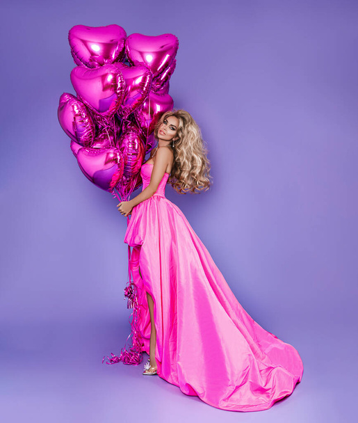 Krásná žena v úžasných dlouhých šatech drží růžové balónky a pózuje na šeříkovém pozadí. Elegantní modelka v šatech. Elegance. Letní galomour móda. - Fotografie, Obrázek