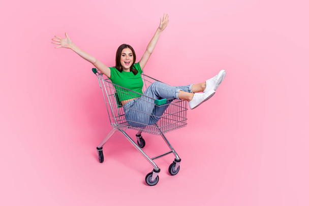 Full length photo of excited cool lady dressed green t-shirt riding shopping cart rising arms hands isolated pink color background. - Фото, зображення