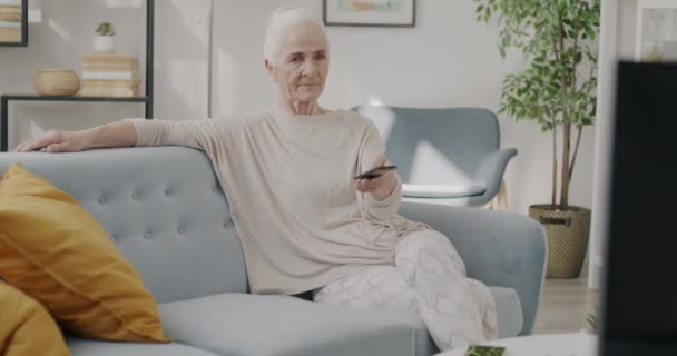 Senior lady watching TV smiling sitting on sofa and using remote control indoors at home. Leisure activity and happy retirement concept. - 映像、動画