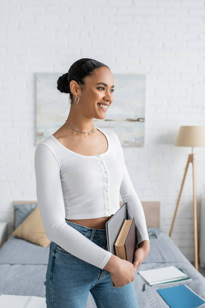 estudiante afroamericano sonriente sosteniendo portátil y reservar en apartamento moderno  - Foto, Imagen