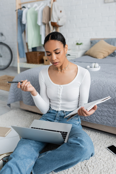 estudiante afroamericano inteligente sosteniendo el cuaderno y la pluma mientras que mira webinar en computadora portátil  - Foto, Imagen