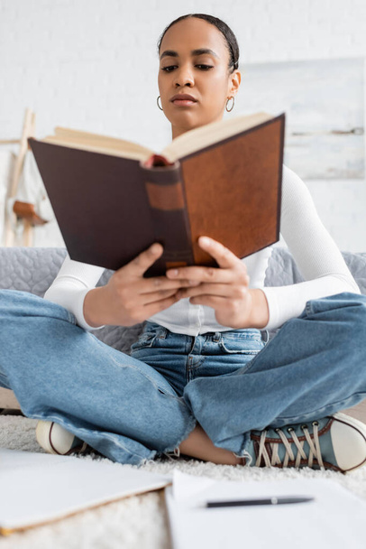 low angle view of young african american student sitting with crossed legs and reading book  - Foto, afbeelding