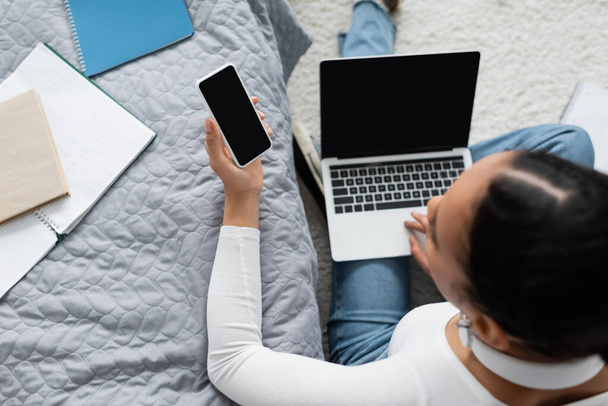 vista dall'alto della donna afro-americana che tiene lo smartphone con schermo bianco vicino al laptop mentre studia da casa  - Foto, immagini