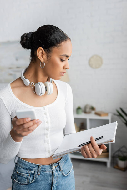 african american student with wireless headphones on neck using smartphone and holding blank notebook in bedroom  - Foto, Imagen