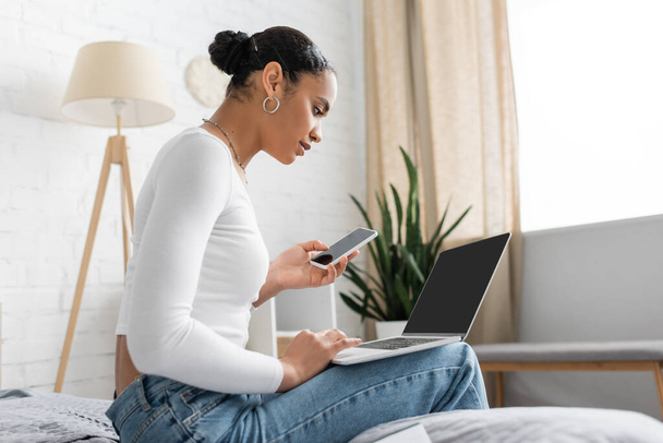молодой африканский студент-американец, держащий смартфон и использующий ноутбук во время обучения онлайн из дома  - Фото, изображение