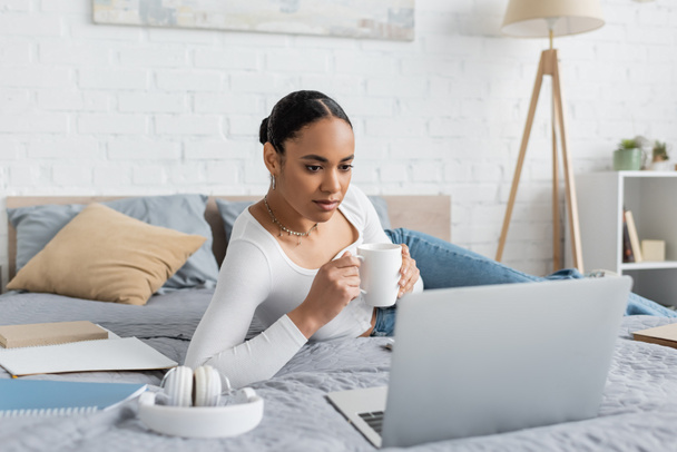 jovem estudante afro-americano segurando xícara de café e olhando para laptop no quarto moderno  - Foto, Imagem