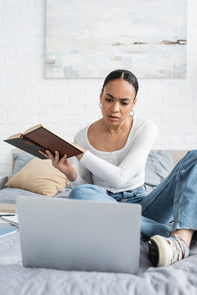 Étudiant afro-américain concentré tenant le livre et regardant l'ordinateur portable sur le lit à la maison  - Photo, image