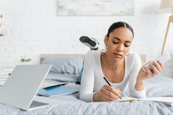 African american student holding smartphone and writing on notebook near laptop on bed  - Photo, Image