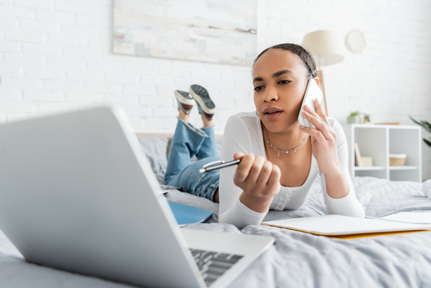 Étudiant afro-américain parlant sur smartphone et regardant un ordinateur portable flou tout en étant couché sur le lit  - Photo, image