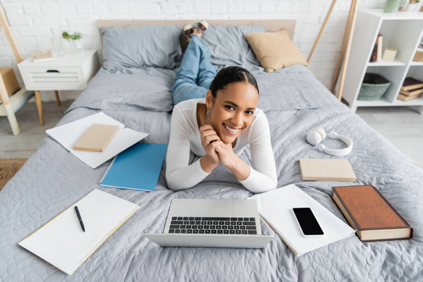 Happy african american student lying near devices and books on bed  - Foto, Imagem