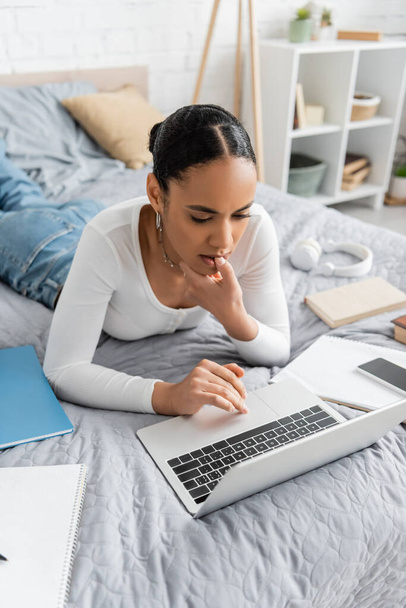 Focused african american student using laptop near notebooks on bed  - 写真・画像