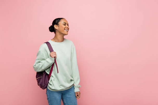 Alegre estudiante afroamericano con mochila mirando hacia otro lado aislado en rosa  - Foto, Imagen