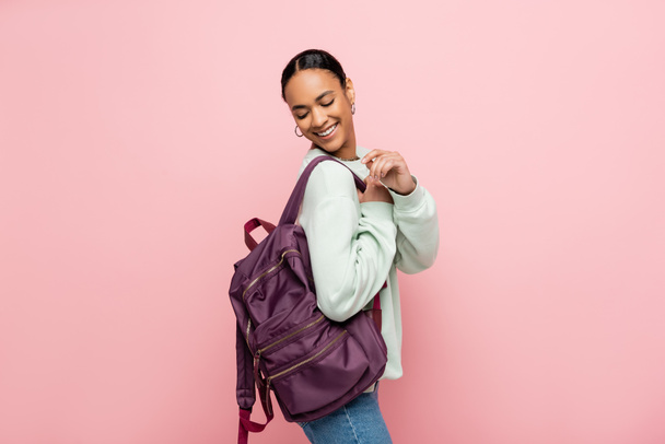 Pretty and smiling african american student looking at backpack isolated on pink  - Foto, Bild