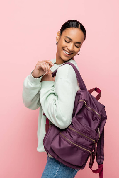 Young african american student in sweatshirt looking at backpack isolated on pink  - Fotó, kép