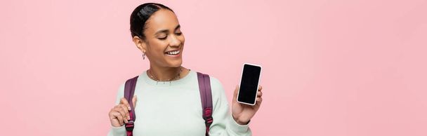 Sırt çantalı gülümseyen Afrikalı Amerikalı öğrenci elinde pembe pankartta izole edilmiş beyaz ekran akıllı telefon tutuyor.  - Fotoğraf, Görsel