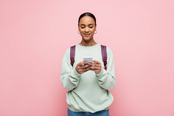 Smiling african american student with backpack typing message on smartphone isolated on pink  - Photo, Image