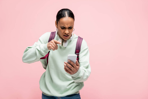 Wütender afrikanisch-amerikanischer Student mit Rucksack und Kopfhörer zeigt vereinzelt auf Smartphone in rosa  - Foto, Bild