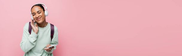 весела афроамериканська жінка в бездротових навушниках тримає смартфон, насолоджуючись музикою ізольовано на рожевому, банер
  - Фото, зображення