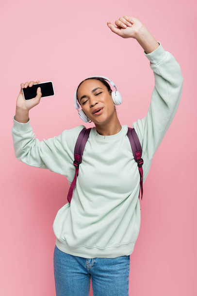 joven estudiante afroamericano en auriculares inalámbricos sosteniendo el teléfono inteligente mientras escucha música y canta en rosa  - Foto, Imagen