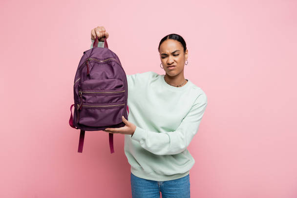 estudiante afroamericano disgustado en sudadera con mochila púrpura aislada en rosa  - Foto, imagen