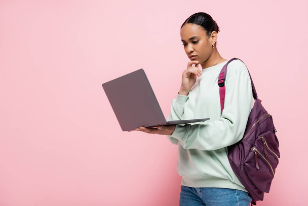 estudante americano africano pensativo com mochila usando laptop em fundo rosa  - Foto, Imagem