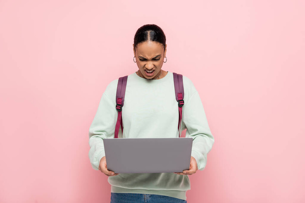 angry african american student with backpack using laptop isolated on pink  - Valokuva, kuva