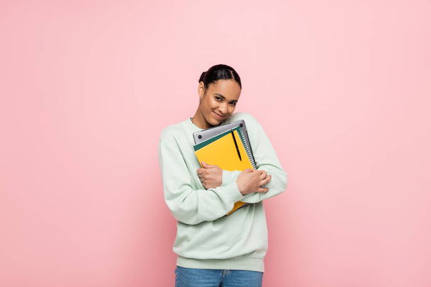 happy african american student in sweatshirt holding laptop and notebooks isolated on pink  - Fotografie, Obrázek