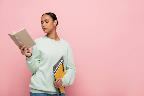 smart african american student in sweatshirt reading book while holding laptop and notebooks isolated on pink  - Photo, Image