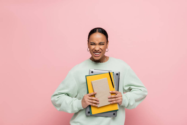 étudiant afro-américain en sweat-shirt tenant ordinateur portable et fournitures d'étude tout en souriant isolé sur rose  - Photo, image