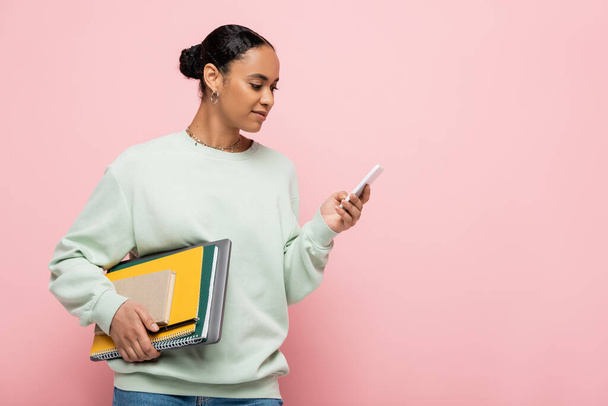 гарненька афроамериканська студентка в сорочці тримає навчальне приладдя і використовує смартфон ізольовано на рожевому
  - Фото, зображення