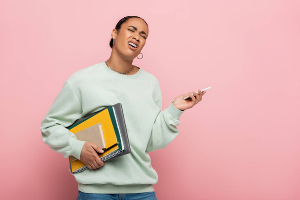 незадоволений афроамериканський студент в сорочці тримає гаджети та навчальні приналежності, одночасно скидаючи ізольовані на рожевий
  - Фото, зображення