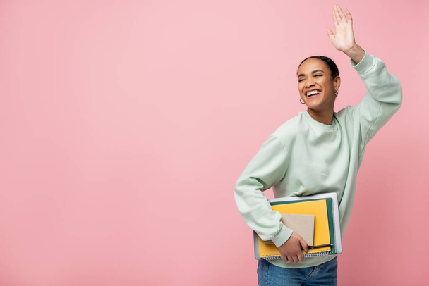 cheerful african american student in sweatshirt holding study supplies and waving hand isolated on pink  - Foto, Bild