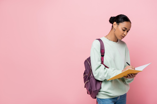 brunette african american student taking notes while holding notebook and pen isolated on pink  - Foto, Imagem