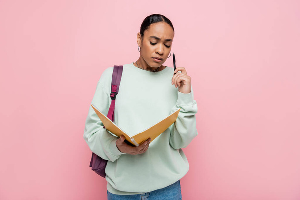 frustrated african american student holding notebook and pen while standing with backpack isolated on pink  - Foto, Imagem