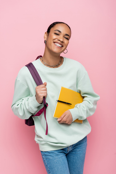cheerful african american student holding notebook and pen while standing with backpack isolated on pink  - Photo, Image