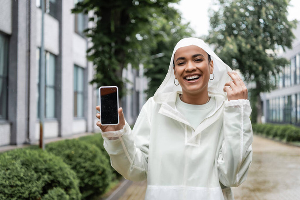 mujer afroamericana feliz en impermeable impermeable con capucha que sostiene el teléfono inteligente con la pantalla en blanco  - Foto, Imagen