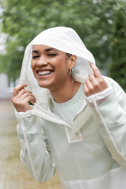 mujer afroamericana llena de alegría en impermeable impermeable ajustando capucha durante la lluvia  - Foto, imagen