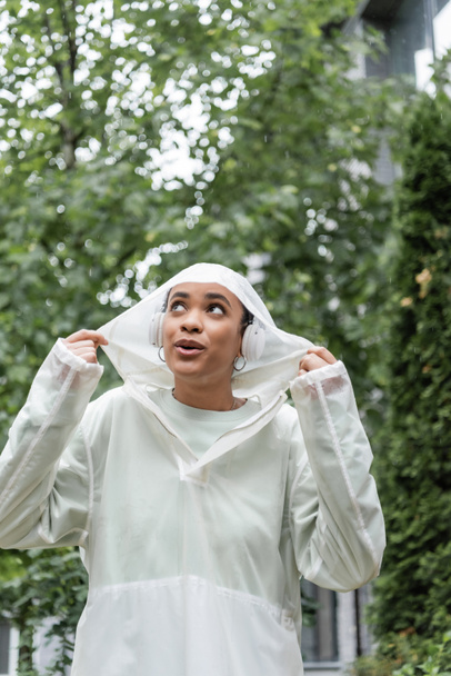 donna afroamericana sorpresa in impermeabile impermeabile e cuffie wireless in piedi sotto la pioggia  - Foto, immagini