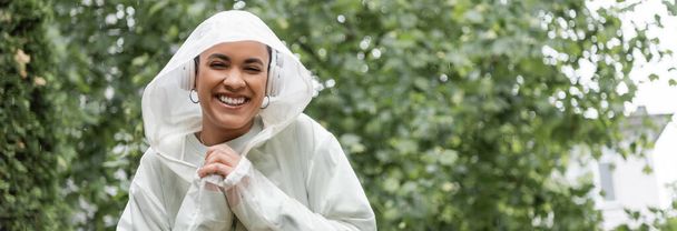 lächelnde Afroamerikanerin in wasserdichtem Regenmantel und kabellosen Kopfhörern, die unter Regen steht, Banner  - Foto, Bild