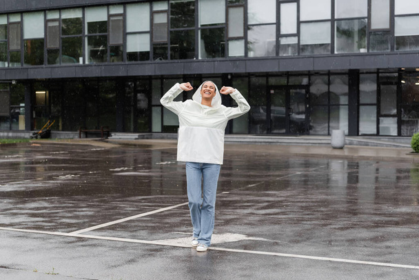 volle Länge einer überglücklichen Afroamerikanerin in wasserdichtem Regenmantel und Jeans, die bei Regen Spaß hat  - Foto, Bild