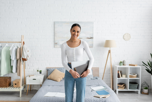 cheerful african american student standing with laptop and book in modern bedroom  - Photo, Image