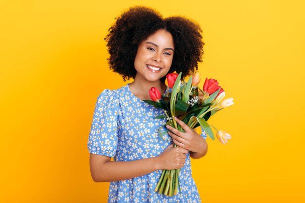 Charming positive african american woman with curly hair, wearing summer blue dress, holding bouquet of colorful tulips, received gift, looks at camera, smiling, stands over isolated orange background - 写真・画像