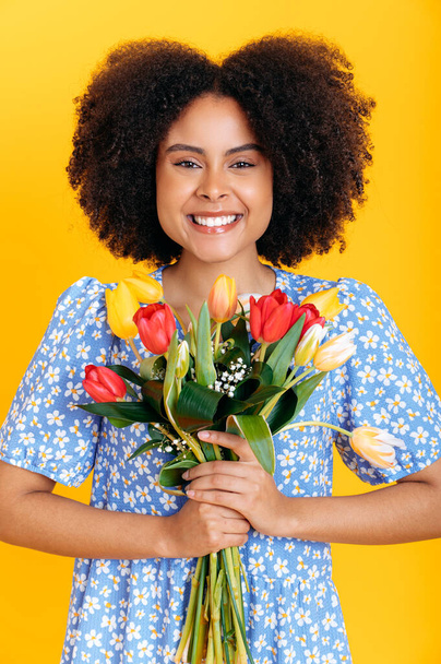 Verticale foto van een mooie positieve Afrikaanse Amerikaanse vrouw met krullend haar, in de zomer blauwe jurk, houdt boeket van kleurrijke tulpen, ontvangen geschenk, kijkt naar de camera, glimlach, geïsoleerde oranje achtergrond - Foto, afbeelding