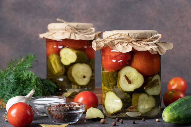 Nakládané okurky s cherry rajčaty ve dvou skleněných nádobách na hnědém pozadí s čerstvými ingrediencemi - Fotografie, Obrázek