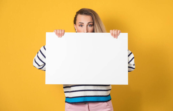 Surprised face of young women behind white board, isolated on yellow background. One girl showing blank empty paper billboard with blank space near her head. Studio shot - Foto, afbeelding