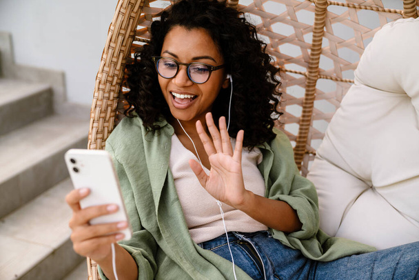 Cheerful young african woman in eyeglasses making video call on smartphone and waving hand while sitting outdoors on rattan hang chair - 写真・画像